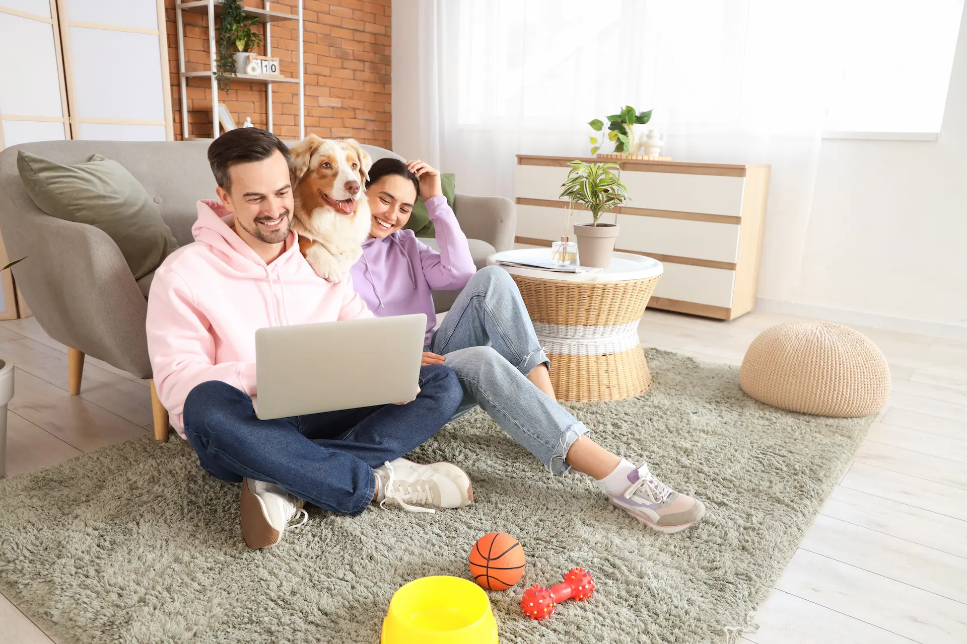 A smiling couple sits on a shaggy rug in a living room, both looking at a laptop held by the man. An Australian Shepherd dog rests its head on the man's shoulder. Pet toys are scattered in the foreground.
