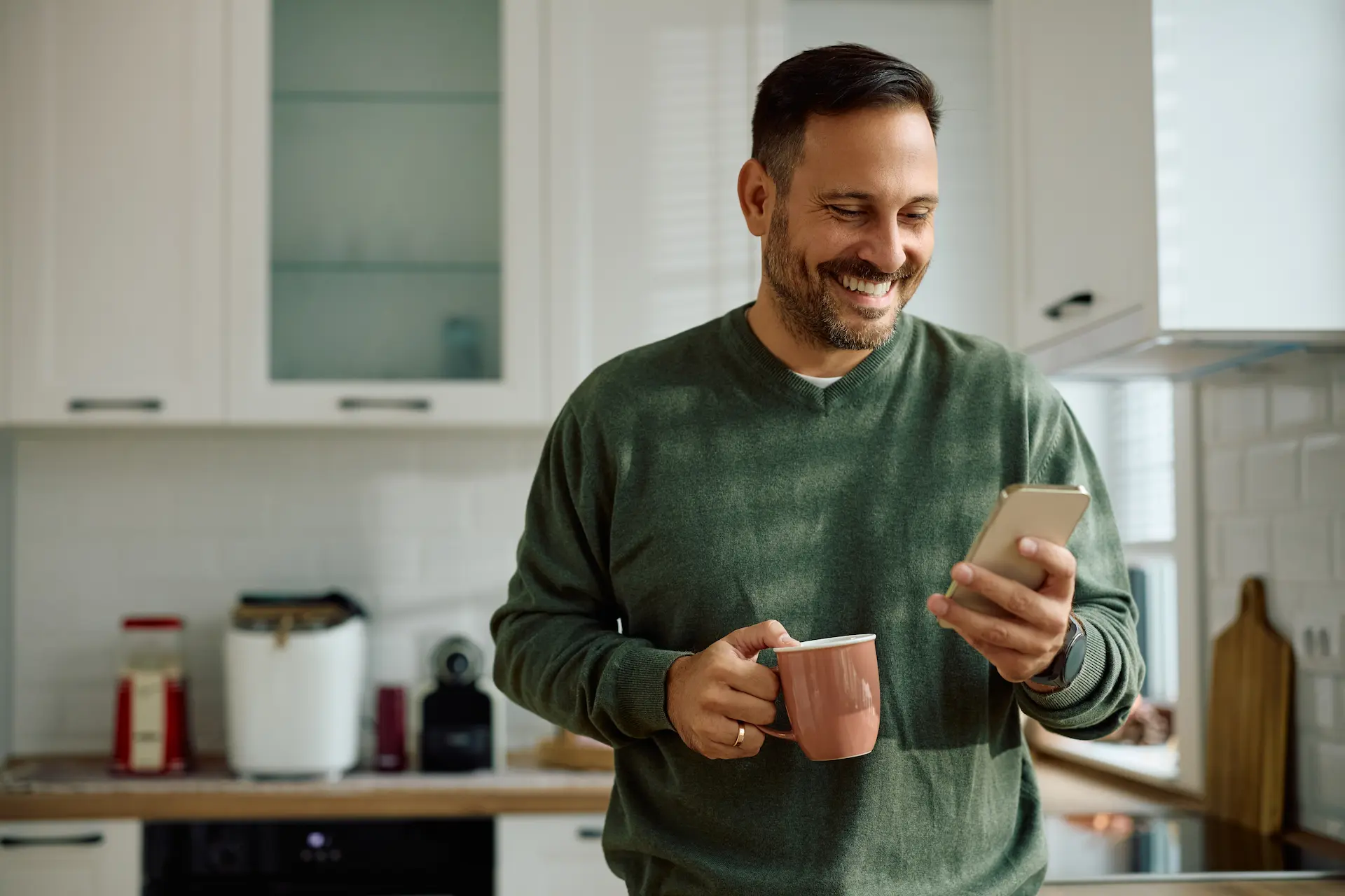 A smiling man in a green sweater holds a coffee mug in one hand and looks at his phone in the other, standing in a kitchen.